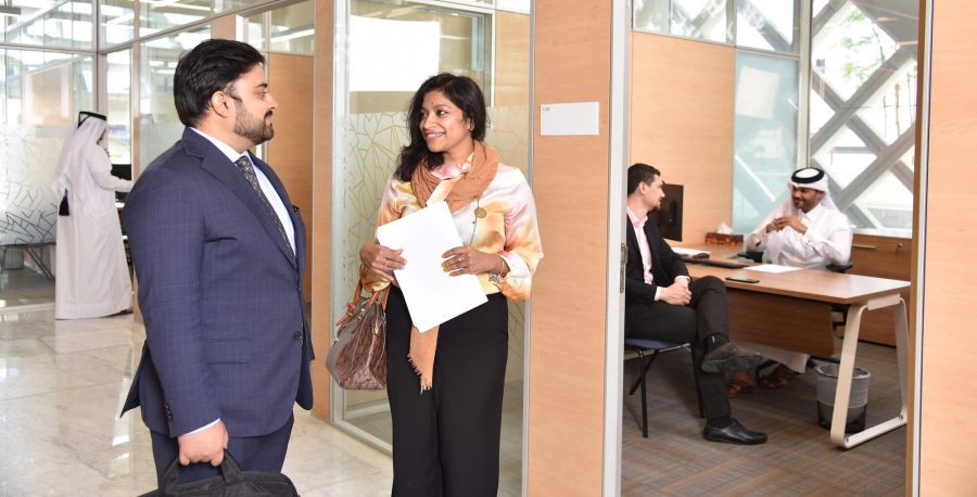 Two Middle-eastern business people, one man and one woman, standing in a bright office, having a happy discussion. Woman is holding paper in her hands. In background other various meetings are taking place