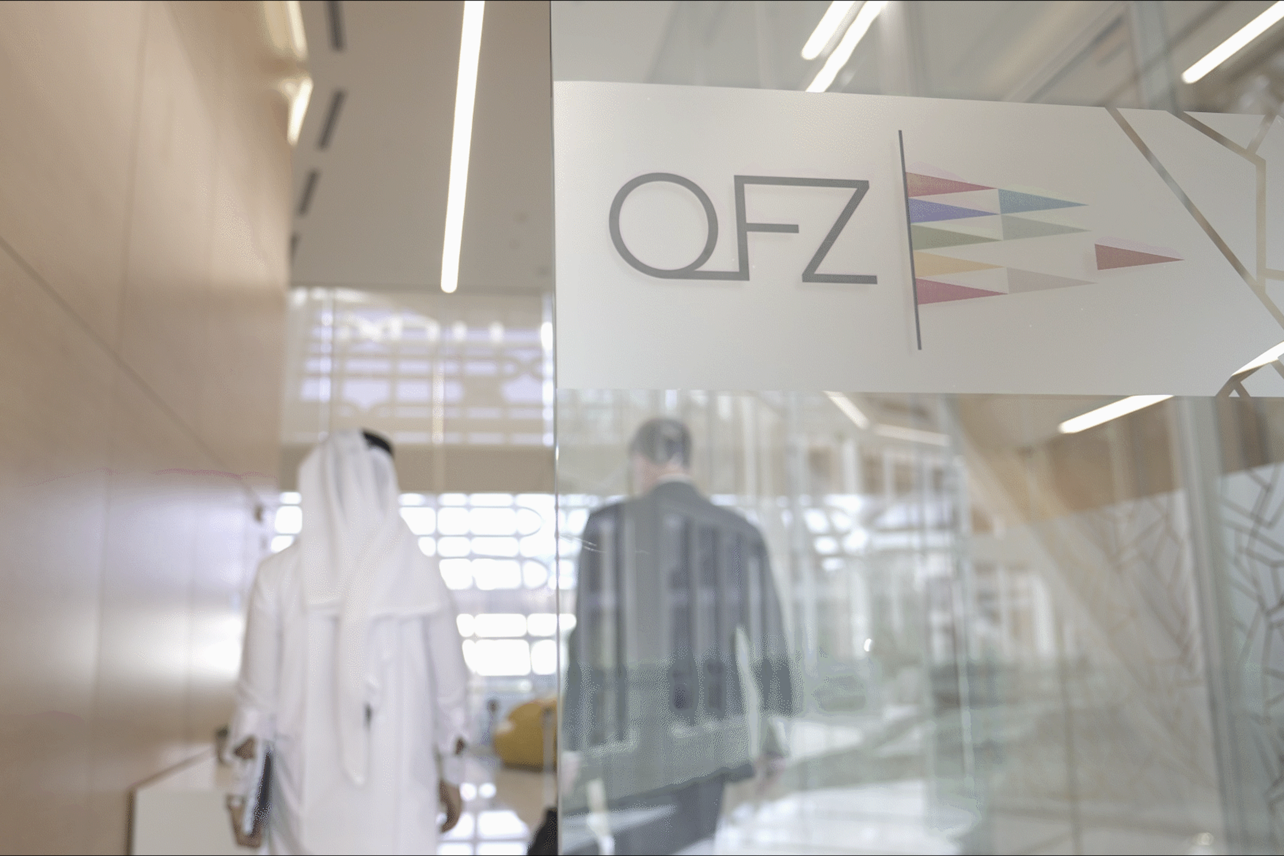 Blurred image of two people in a office, behind opaque glass wall with Qatar Free Zones logo
