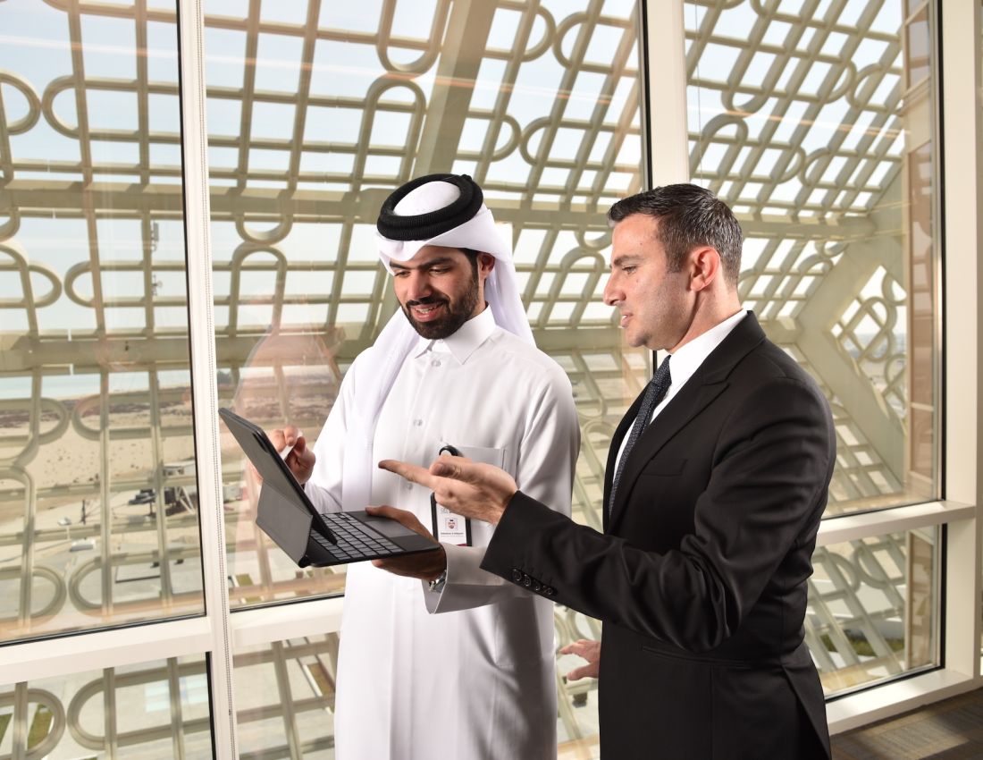 Two businessmen, one in a suit and one in traditional Arabic clothing, standing together discussing while pointing at a computer screen