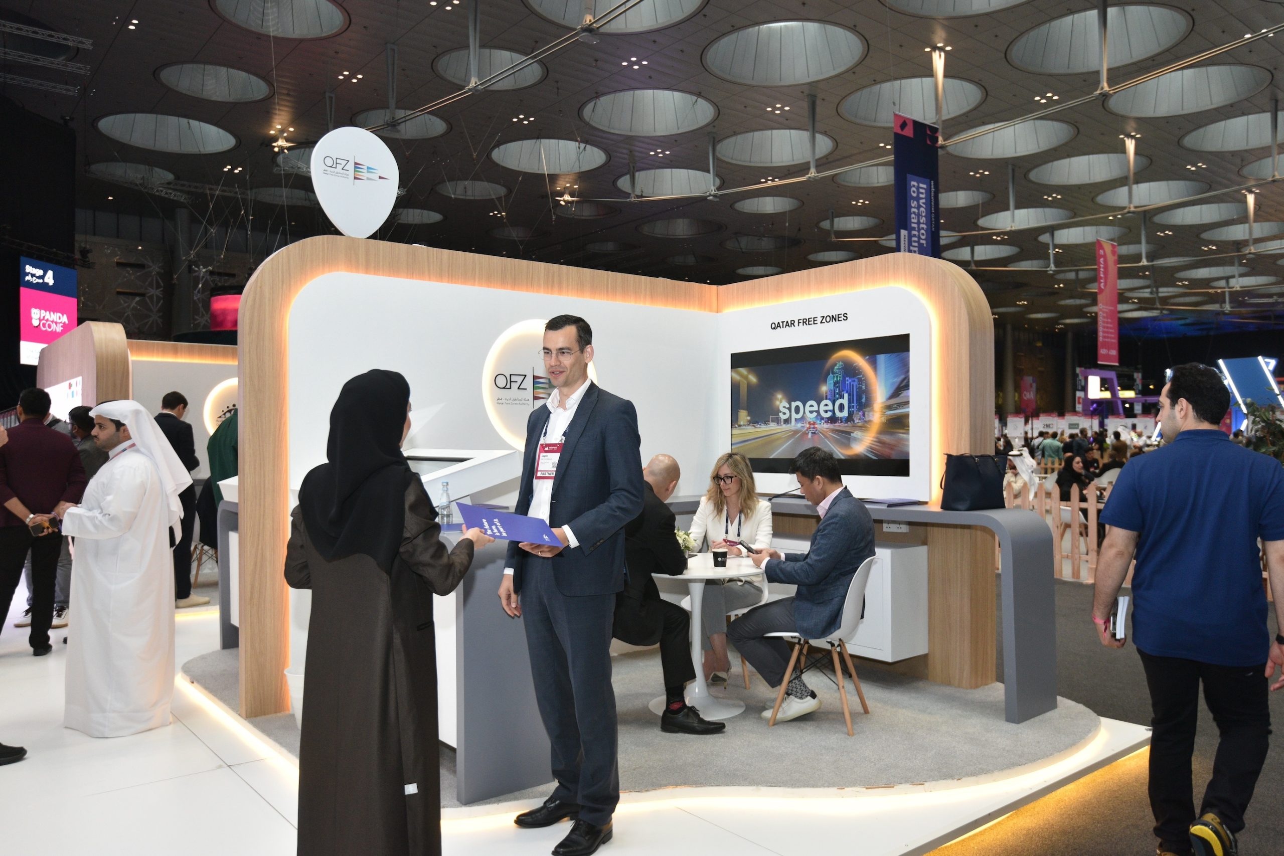 Lively business discussion between a man and a woman, one in traditional Arabic clothing and one in a suit in front of the QFZ booth at Web Summit Qatar 2024
